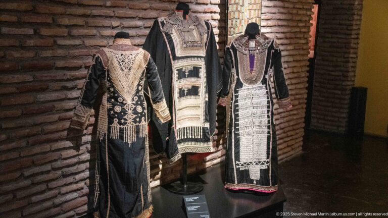 Three dark traditional robes on mannequins with cream woven panels, embroidery, fringe, and colored trim.