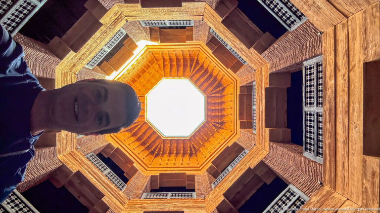 Steep upward view through MAP Marrakech’s octagonal light well, with terracotta brick, timber galleries, screened windows, and bright sky.