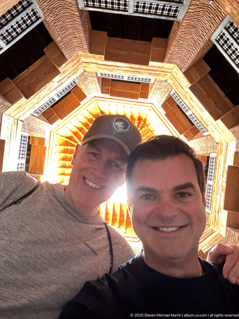 Two smiling visitors pose beneath an octagonal roof opening framed by brick walls, timber beams, and screened windows.