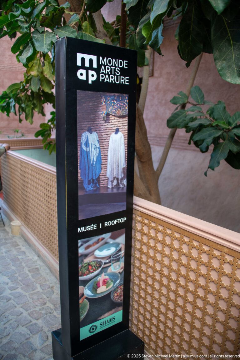 Black Monde des Arts de la Parure sign beside greenery, with garment images and directions for the museum and rooftop.