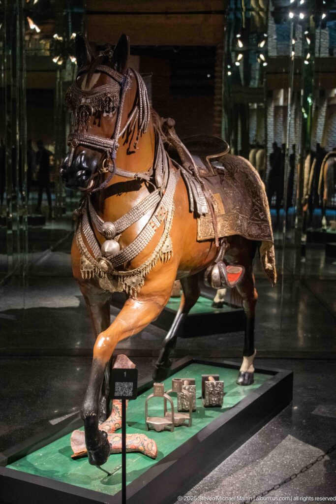 Life-size horse display wearing an ornate saddle, bridle, chest straps, and decorative fittings in a museum case.