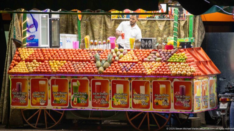 Fruit-juice vendor behind a wheeled stall stacked with oranges, pomegranates, bananas, and other produce under bright lights.
