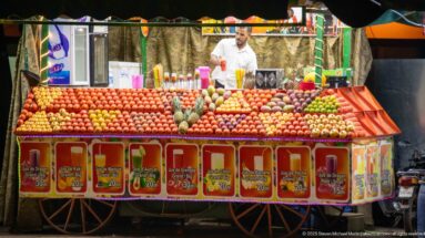Fruit-juice vendor behind a wheeled stall stacked with oranges, pomegranates, bananas, and other produce under bright lights.