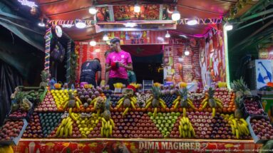 Food stall No. 15 with two workers behind a symmetrical display of bananas, apples, citrus, and other produce.