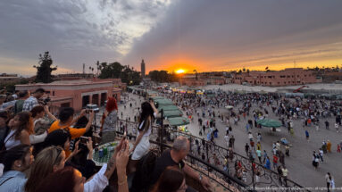Visitors at a rooftop railing photographing sunset over the crowded Djemaa El Fna square.