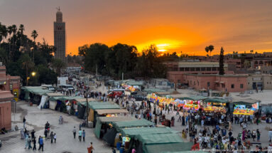 Busy Djemaa El Fna market stalls and pedestrians beneath an orange sunset and silhouetted palms.