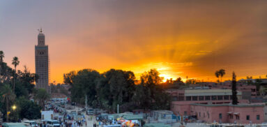 Koutoubia Mosque minaret above palms and streets beneath a radiant orange sunset.