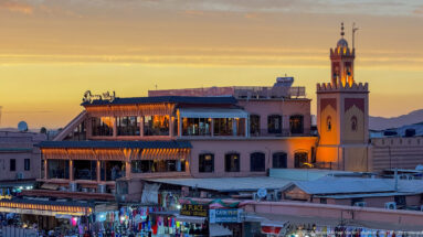 Illuminated square minaret beside rooftop restaurants and bright market storefronts at dusk.