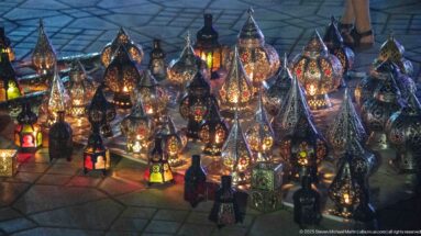 Cluster of glowing pierced-metal lanterns in varied shapes, some fitted with colored glass panels, displayed on the ground.