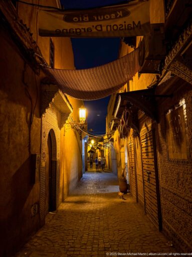 Quiet cobbled medina lane at night with closed shopfronts, fabric awnings, and warm wall lamps.