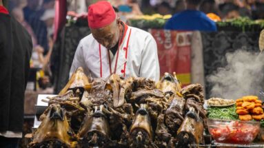 Cook in white coat and red cap behind a large pile of prepared sheep heads at a steaming food stall.