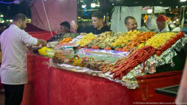 Vendor beside a food counter packed with sausages, skewers, fried foods, vegetables, and condiments under bright market lights.