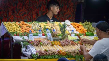 Young vendor behind a brightly arranged food stall with skewers, seafood, fried items, herbs, citrus, and pineapples.