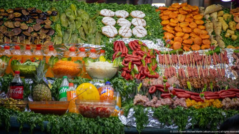 Close view of a market food display with skewers, sausages, greens, fried foods, vegetables, sauces, and bottled drinks.