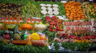 Close view of a market food display with skewers, sausages, greens, fried foods, vegetables, sauces, and bottled drinks.