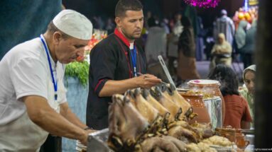 Two food-stall workers preparing dishes beside a foreground stack of cooked sheep heads and a large earthenware pot.