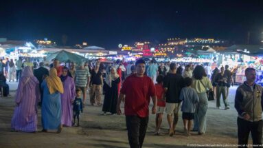 Crowds walking through Jemaa el-Fna at night with brightly lit market stalls stretching across the square behind them.