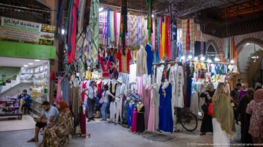 Covered souk passage lined with robes, dresses, scarves, and colorful hanging fabrics as shoppers walk through the arcade.
