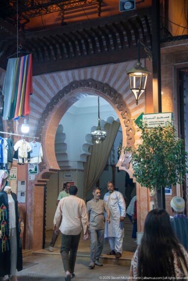 Pedestrians pass beneath a broad brick-trimmed horseshoe arch with hanging lantern and market signs in a Marrakech souk.