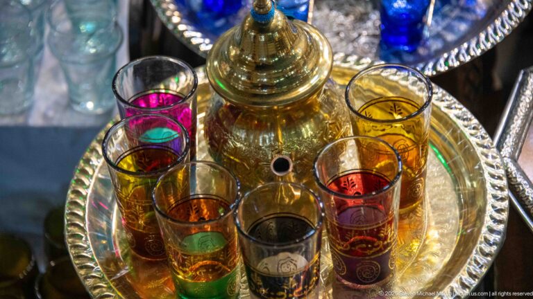 Polished teapot surrounded by slender red, green, amber, and clear tea glasses on a round metal tray.