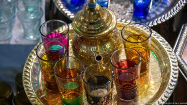 Polished teapot surrounded by slender red, green, amber, and clear tea glasses on a round metal tray.