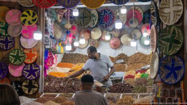 Vendor reaching over bins of dried fruits, nuts, legumes, and spices beneath brightly patterned woven baskets.