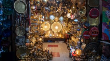 Interior of a crowded souk shop filled with glowing pierced lamps, hanging lanterns, trays, vessels, and decorative metalware.