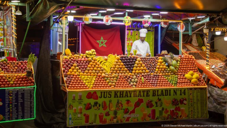 Fruit-juice stall marked Jus Khayrate Bladi N°14, with Moroccan flag, vendor, and patterned rows of fresh fruit.