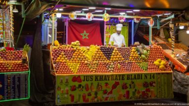 Fruit-juice stall marked Jus Khayrate Bladi N°14, with Moroccan flag, vendor, and patterned rows of fresh fruit.