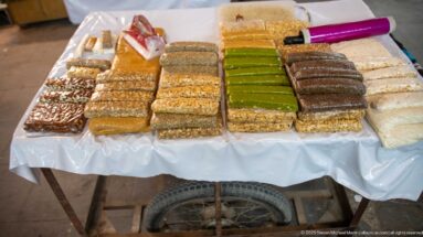 Rows of rectangular nut, sesame, pistachio-colored, and nougat-like sweets arranged on a market table.