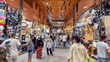 Busy covered souq passage lined with shops, merchandise, and pedestrians beneath a slatted roof.