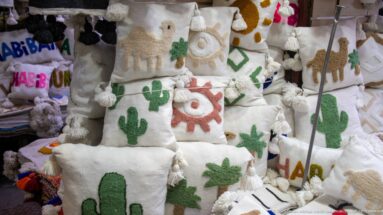 Stacked white woven souvenir bags with colorful camel, cactus, eye, and palm motifs.