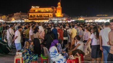 Dense nighttime crowd gathered around street activity in Jemaa el-Fna, with seated women in the foreground and illuminated buildings behind.