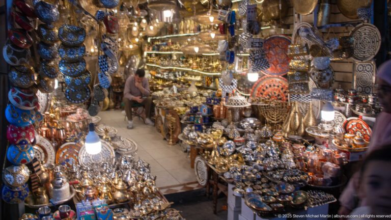 Narrow shop glowing with lamps and packed with trays, teapots, bowls, and decorative metalware.