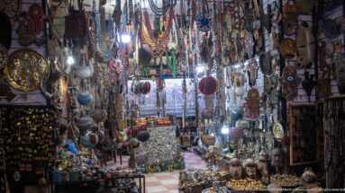 Narrow souvenir shop crowded with hanging necklaces, charms, masks, and small decorative objects.