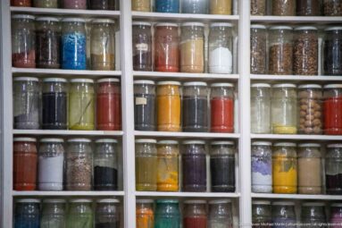 Grid of glass jars filled with colored powders, dried herbs, seeds, and other market ingredients.