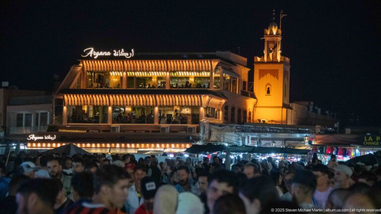 Illuminated Café Restaurant Argana above a packed nighttime crowd in Jemaa el-Fna, with striped awnings and rooftop terraces.