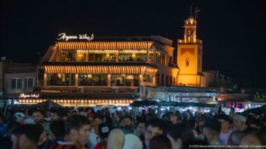 Illuminated Café Restaurant Argana above a packed nighttime crowd in Jemaa el-Fna, with striped awnings and rooftop terraces.