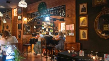 Warm restaurant interior with diners, exposed brick, framed art, dark walls, and carved wood trim.