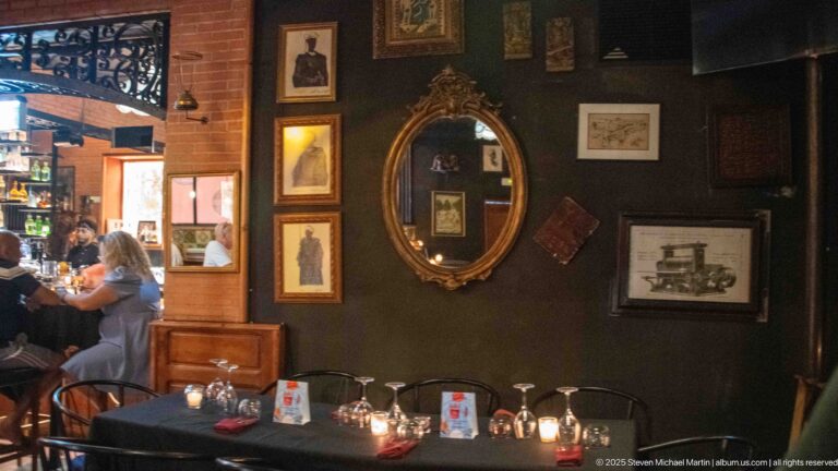 Dark restaurant wall with oval gilded mirror, framed prints, candlelit tables, and wood furnishings.