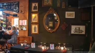 Dark restaurant wall with oval gilded mirror, framed prints, candlelit tables, and wood furnishings.