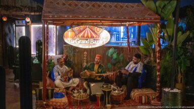 Three seated musicians performing beneath a Le Grand Bazar sign in a richly decorated restaurant setting.