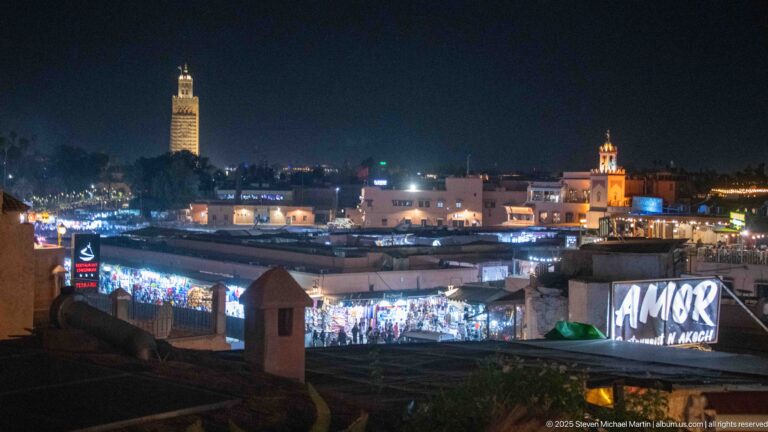 Night view across low medina rooftops toward illuminated minarets and brightly lit market buildings.