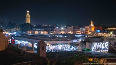 Night view across low medina rooftops toward illuminated minarets and brightly lit market buildings.