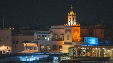 Illuminated red-and-cream minaret rising above restaurant terraces and low rooftops at night.