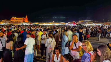 Wide nighttime view of Jemaa el-Fna filled with crowds, with a long band of bright market stalls across the background.
