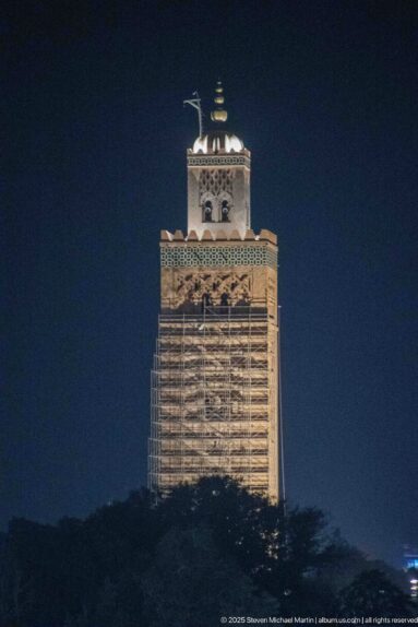 Distant night view of the square Koutoubia minaret rising above dark trees with its upper section illuminated.