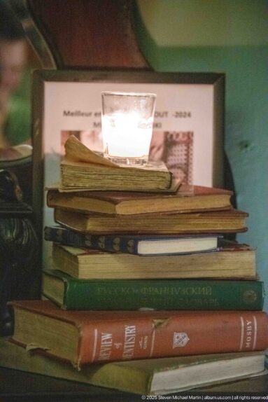 Stack of worn books topped with a glowing glass candle holder against a green wall.