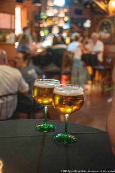 Two stemmed amber drinks on a dark table with blurred diners and lights behind.