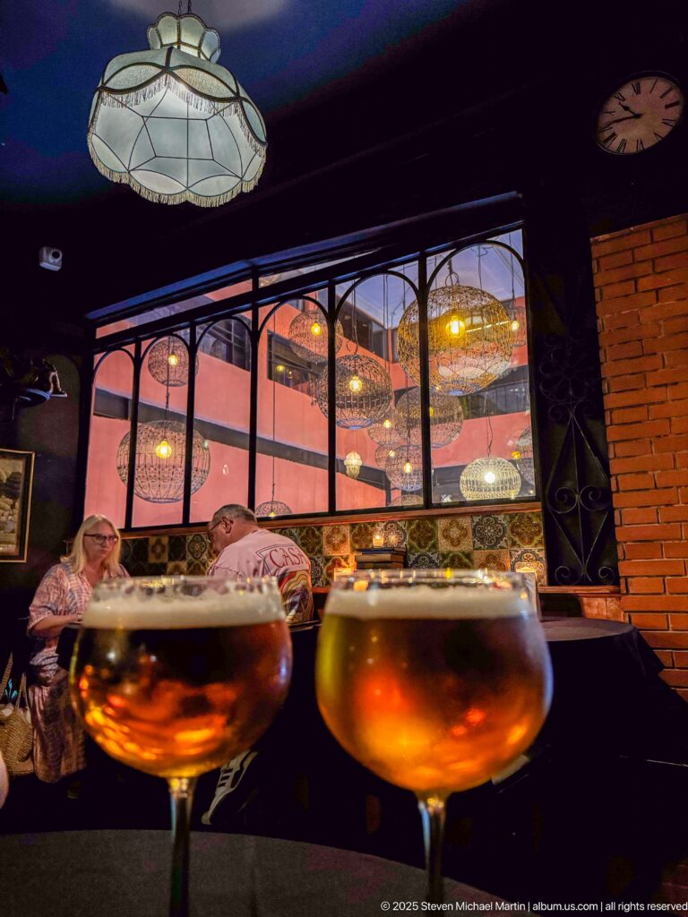 Two amber drinks in front of a decorated dining room with hanging lamps and seated patrons.